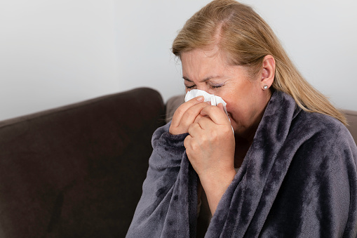 Portrait of a sick senior woman with flu blowing her nose on couch in living room.
