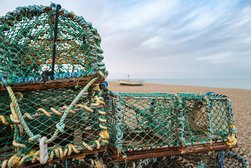Shallow focus of stacked lobster pots and cages seen on an empty Suffolk beach. A distant small boat can be seen on the shingle beach front.