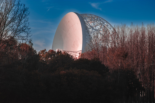 An aerial photograph of Jodrell Bank, Cheshire, England. The photograph was produced on a bright autumn afternoon with clear blue skies. The photograph shows the top of autumnal trees overlooked by the Jodrell Bank telescope.