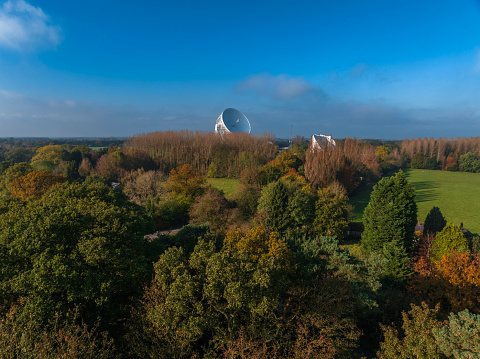 An aerial photograph of Jodrell Bank, Cheshire, England. The photograph was produced on a bright autumn afternoon with clear blue skies. The photograph shows autumnal trees meeting the bright blue sky on the horizon. Also sitting on the horizon is a large radio telescope.