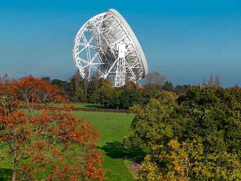 An aerial photograph of Jodrell Bank, Cheshire, England. The photograph was produced on a bright autumn afternoon with clear blue skies. The photograph shows a radio telescope surrounded by autumnal trees and the beautiful Cheshire landscape.