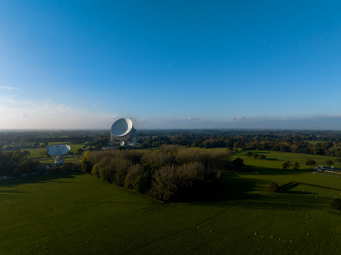 An aerial photograph of Jodrell Bank, Cheshire, England. The photograph was produced on a late autumn afternoon with clear blue skies. The photograph shows three radio telescopes situated amongst autumnal trees and the beautiful Cheshire countryside.