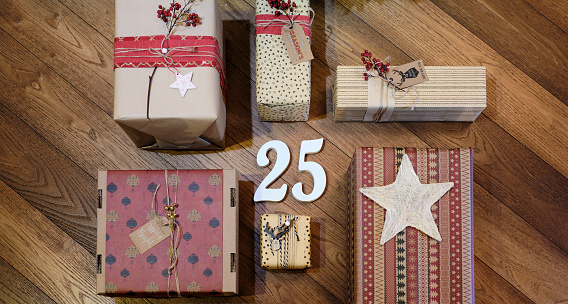 Top view of Christmas packages wrapped with gift cards decorated with Christmas motifs. In the center, the number 25 wishing you a Merry Christmas. In the background a wooden floor with warm colors.