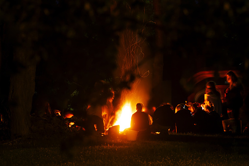 People around a bonfire at night. Figure silhouettes. Unrecognizable. Warm atmosphere. Group singing. Camping. Free time activity.