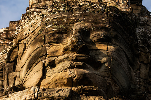 A huge face carved in stone, seen from below, low angle shot, illuminated by the afternoon light at Bayon temple, Angkor, Cambodia.