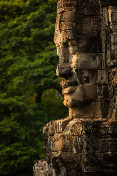 ogromna twarz wyrzeźbiona w kamieniu, czuwa nad zwiedzającymi świątynię bayon, angkor, kambodża, w tle otacza ją ściana zielonej dżungli - wat angkor thom zdjęcia i obrazy z banku zdjęć