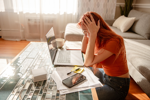 Beautiful young girl with red hair and glasses is working from home, multitasking. She's resolving work-related issues - Stock Photo