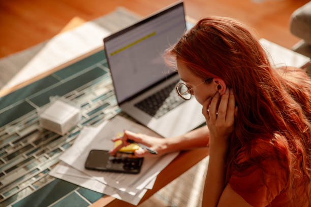 istock Red-haired girl with glasses, using a calculator and paying bills online - Multitasking 1763098921