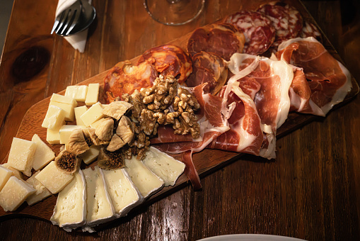 There are various cheeses, ham, and sausages on a wooden platter.
