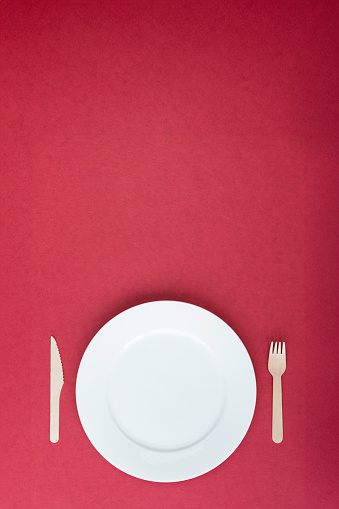 Directly above view of empty white plate and wooden cutlery on red background. Representing recycle and sustainability concepts in food industry.