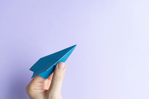 Hand is holding blue colored paper plane in front of purple colored background.