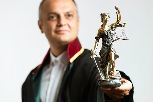 Male lawyer is showing Lady Justice sculpture  to camera in front of gray background and is looking up.
