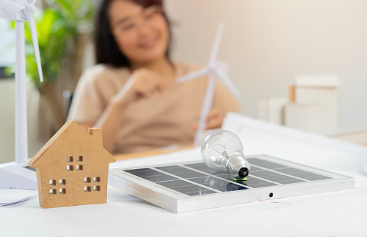 Close up of solar panel, light bulb and house wood model on the table with blurred background of girl holding wind turbine. Concept clean energy and saving power in nature.