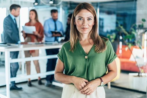Portrait of a young business woman in the modern office, and a team working behind her. Successful businesswoman standing in creative office and looking at camera.