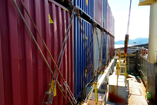 Row of lashed containers in a line with bars and twist locks situated in forward of container vessel with cranes. Behind is hill of Brazilian coast.