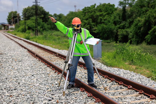 Engineer asian male wearing green uniform and orange hard hat use theodolite equipment and holdint data paper surveying construction worker on Railway site. Repair and maintenance raiway lines.