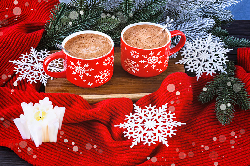 Christmas composition with two red coffee cups, snowflakes, lollipop, burning white candle, Christmas tree branches, red scarf on snowy background.