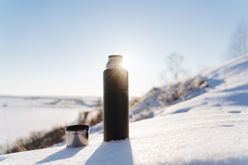 Sunlight glints with rays against the blue sky. A thermos of hot tea stands in the snow. Winter landscape, hiking in the forest, camping equipment. High quality photo