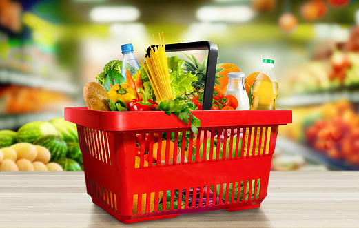 Food and groceries in red shopping basket on wood table on blurred supermarket aisle background