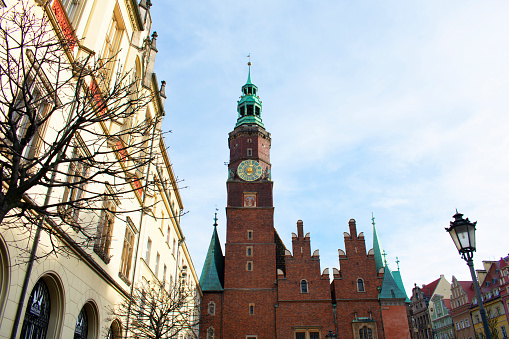 Tall tower of old Catholic church made of red brick, with green towers, sculptures and green clock. Old buildings around, lantern, tree branches without leaves. Old town. Poland, Wroclaw, January 2023