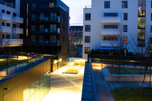 Night view of a modern residential complex with a long alley, lawns and trees. Glass elements in balconies and walls. Night lighting with yellow and blue lights. Realty. Wroclaw, Poland, January 2023.