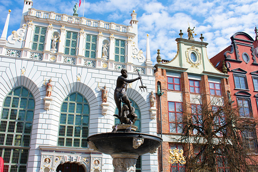 View of the old facades of white and pink buildings with high windows, arches, statues and decorative elements. Central square with the statue of Neptune. Blue sky. Poland, Gdansk, April 2023