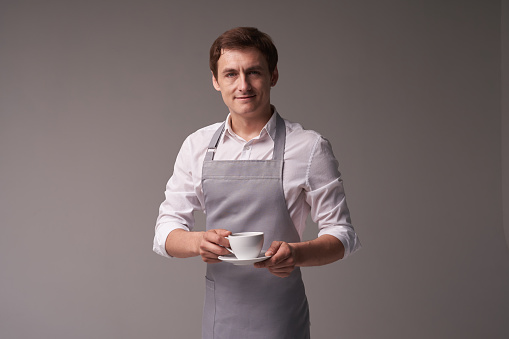 The positive male waiter on a grey background. The barista holds a coffee drink for the customer. Portrait of a successful young man. Layout with copy space. High quality photo