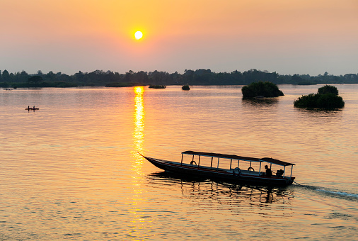 Silhouette of river craft,drifting across the calm,peaceful waters of the Mekong,through rays of golden light reflected on the surface of the river water, by the setting sun.