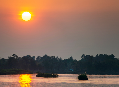 Si Phan Don.Silhouettes of small islands and trees, across the calm,peaceful waters of the Mekong,the fat setting sun hanging in the sky.Shining golden light reflected on the water, in southern Laos.