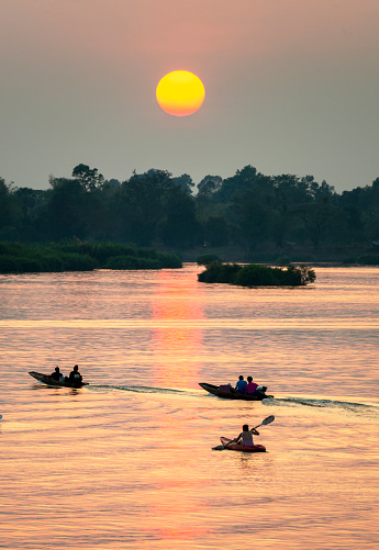 Silhouettes of human figures in small,narrow boats,drifting across the calm,peaceful waters of the Mekong,through rays of golden light reflected on the water, from the setting sun,in southern Laos.