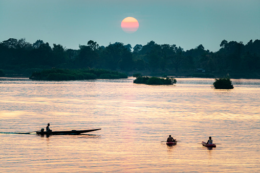 Silhouettes of human figures in small boats,drifting across the calm,peaceful waters of the Mekong,through rays of golden light reflected on the water, from the setting sun,in southern Laos.