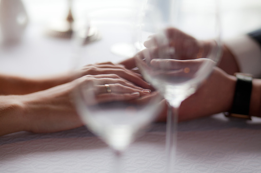 An intimate close-up view of two individuals' hands resting on a white tablecloth, one wearing a golden ring, with wine glasses subtly in focus in the foreground. The soft lighting accentuates the intricate details of the hands and brings out the tranquil ambiance of the moment.