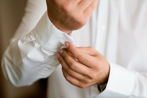 A close-up view of a young man's hands skillfully fastening a silver cufflink to his crisp white shirt's cuff, preparing for a formal occasion. The natural lighting accentuates the fine details of the fabric and the shimmer of the cufflink, conveying a sense of elegance and anticipation.