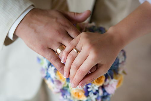 A touching close-up capturing a moment of intimacy and connection. It showcases two hands, with the focus on the wedding rings,against the background of a bouquet of flowers. The soft pastel colors of the bride's floral bouquet provide a serene backdrop, encapsulating the essence of love, commitment, and the beginning of a lifelong journey together.