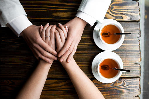 A tender moment between two individuals, hands lovingly intertwined atop a rustic wooden table, signaling trust and affection. Nearby, two cups of warm tea give off a comforting vibe, denoting a relaxed and intimate setting. The top-down angle of the photograph accentuates the focus on the hands and their significance.