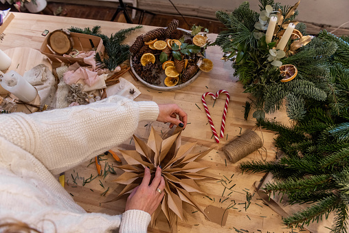 Faceless girl is adjusting star made of craft paper made with own hands on wooden table. Natural home decorations for Christmas. Around star are candles, fir branches, tools, balloons, dried oranges