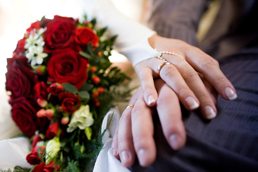 A beautifully intimate moment at a wedding ceremony, this close-up image captures a bride and groom's hands gently entwined. With the bride's pristine white gown and delicate pearl bracelet on one side and the groom's sharp gray suit on the other, the center focus is on their newly adorned wedding rings. A bouquet of deep red roses with hints of green foliage offers a rich backdrop, further emphasizing the central theme of love and unity.