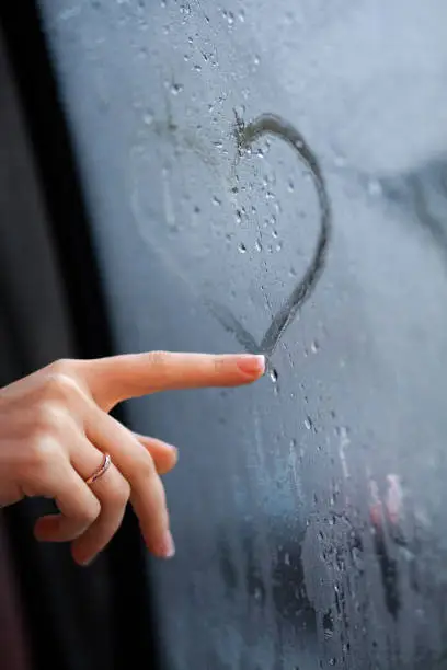 A poignant moment is captured as a woman's hand, adorned with a delicate ring, gently traces a heart shape on a misted windowpane. The image exudes a sense of melancholy mixed with hope, as droplets of rain gather outside, and the heart, only partially formed, conveys the ephemeral nature of love. The focus remains sharp on the fingertip and the immediate area of the heart, emphasizing the act of drawing, while the surrounding window and external environment are artfully blurred.
