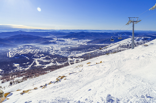 Winter nature panorama with Sheregesh ski resort in Altai, Russia, picturesque nature, sun flare on blue sky, white snow slopes and ski track, top view with range mountains, forest, aerial perspective