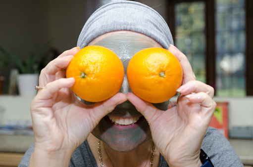 Headshot of mature woman with facial beauty mask and holding two orange in front of her eyes