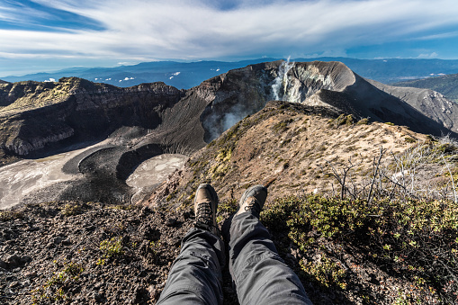 man in hiking boots watching a volcanic landscape with rocky craters and smoking arids on a sunny morning