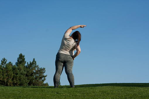 overweight middle-aged woman does exercises in park standing on grass, blue sky background, back view. fat woman tilts body to side. Healthy lifestyle, body positivity, Pilates outdoors in summer
