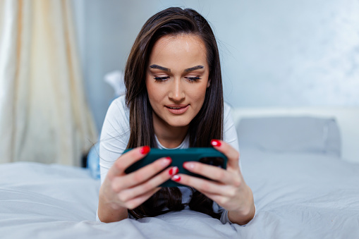 Young Caucasian female student relaxing on her bed at home, while watching a movie on her mobile phone.