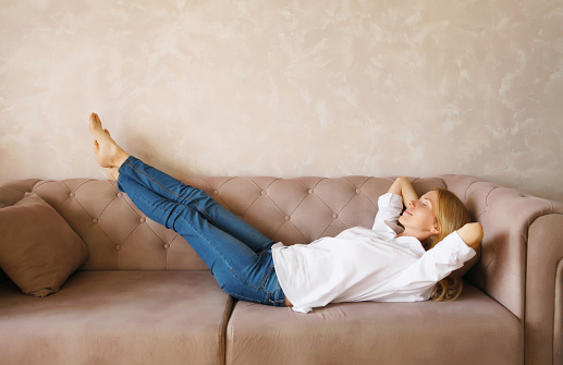 Relaxed caucasian young woman resting with closed eyes and folded hands behind the head while lying on couch in the living room at home