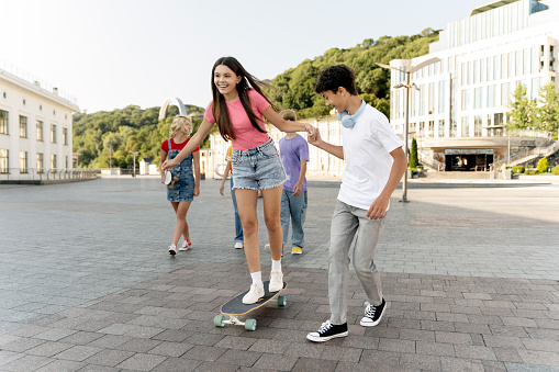 Portrait of smiling teenage friends riding on skateboard on urban street. Group of happy multiracial boys and girls having fun together. School vacation, summer, positive lifestyle concept
