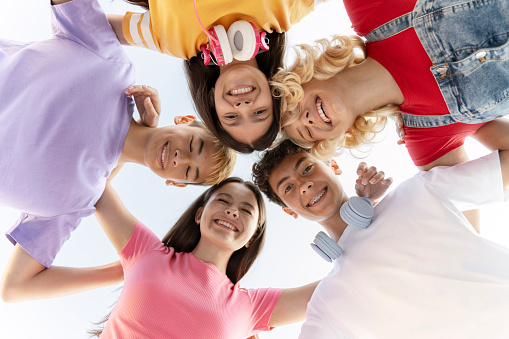 A large group of ethnically diverse teens, lay in the grass in a circle as they pose for a portrait.  They are each dressed casually and are smiling in this aerial view.