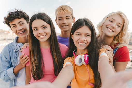 Portrait of smiling multiracial teenage friends taking selfie, looking at camera on urban street. Young influencer recording video standing together with friends. Summer concept