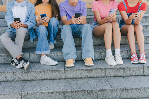 Cropped picture of group teenagers holding mobile phones watching video, communication online, chatting sitting on the street. Technology, social media concept