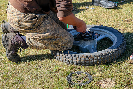 A nomadic herder in the Altai mountains, wearing combat trousers & work boots, kneeling on a motorbike wheel, that he is repairing on an area of grass. Using a handheld wrench he is tightening wheel bolts.