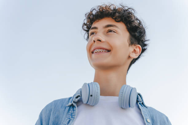 Smiling curly haired teenager with braces wearing headphones looking away standing on the street Portrait of smiling curly haired teenager with braces wearing headphones looking away standing on the street. Inspiration, technology concept teenager boy haircuts stock pictures, royalty-free photos & images